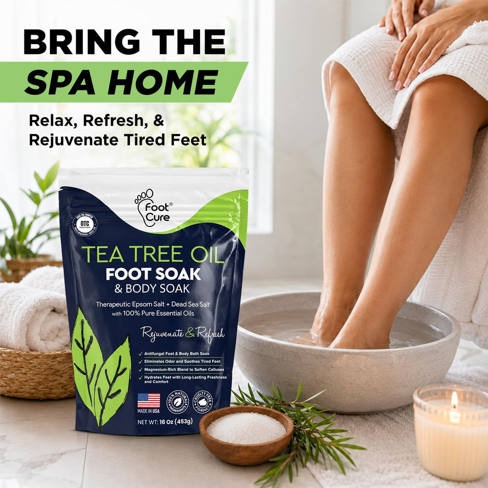 A bag of Root Cure Tea Tree Oil Foot Soak and Body Soak stands on a bathroom floor next to a person sitting in a spa-style setting with their legs in a foot bath bowl. The scene includes towels, a candle, and decorative plants, with promotional text at the top reading 'BRING THE SPA HOME'.