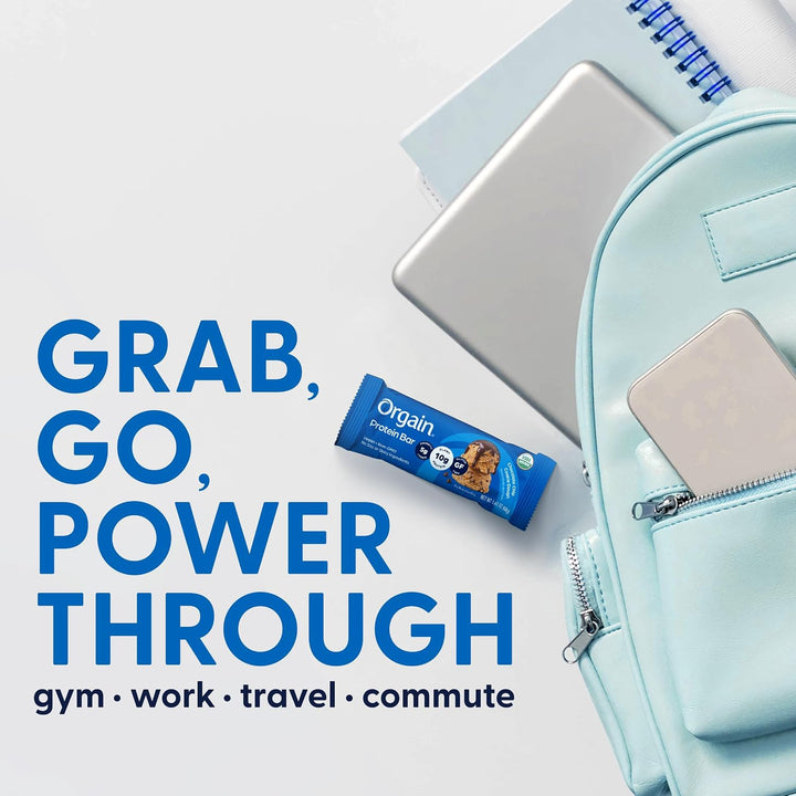 A light blue backpack with a phone in its front pocket, a silver laptop, a notebook, and an Orgain Protein bar, arranged on a white background with the text 'GRAB, GO, POWER THROUGH' and 'gym • work • travel • commute'.